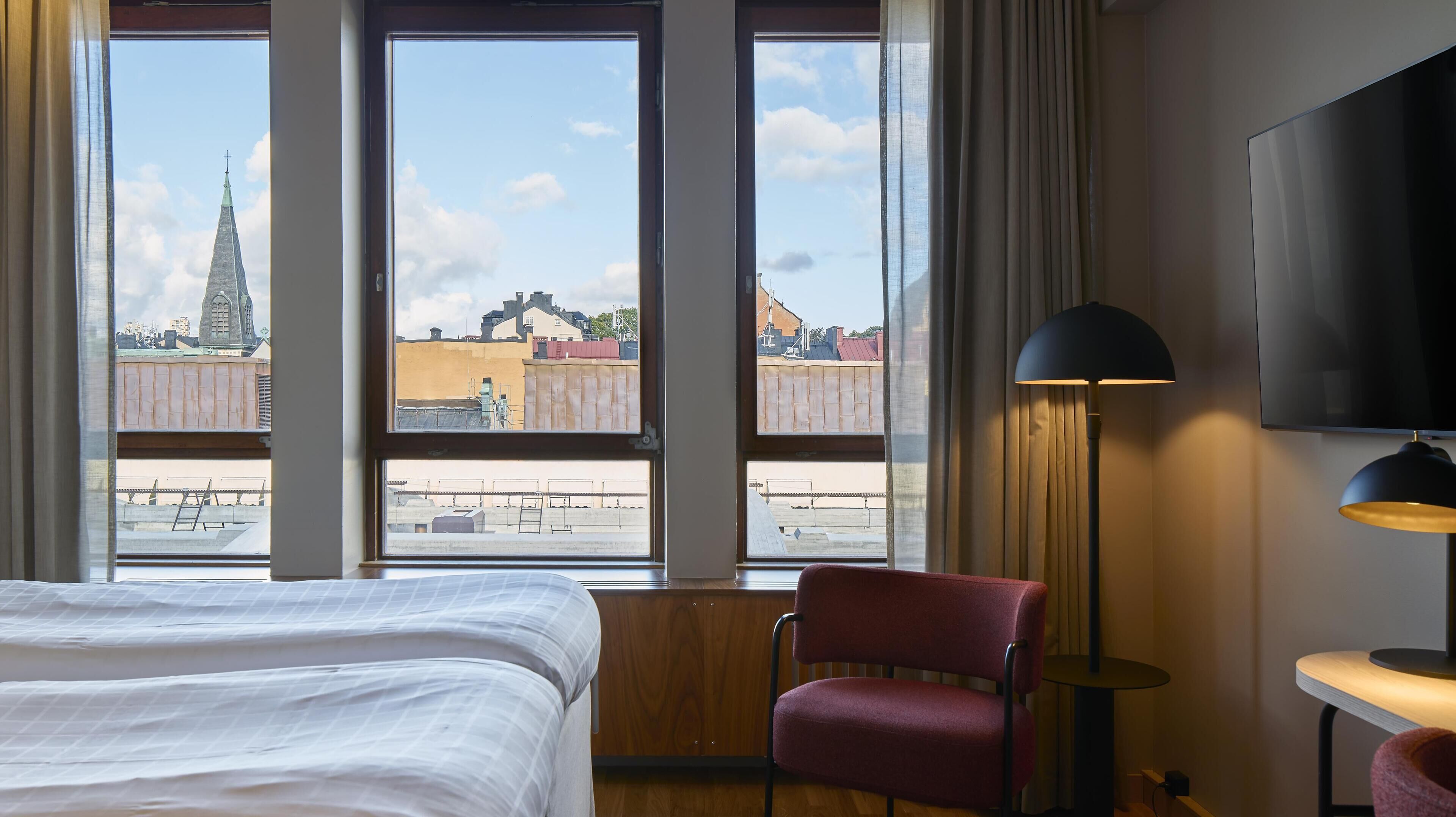A hotel room with a large bed, a red chair, a desk, a lamp, and a TV.