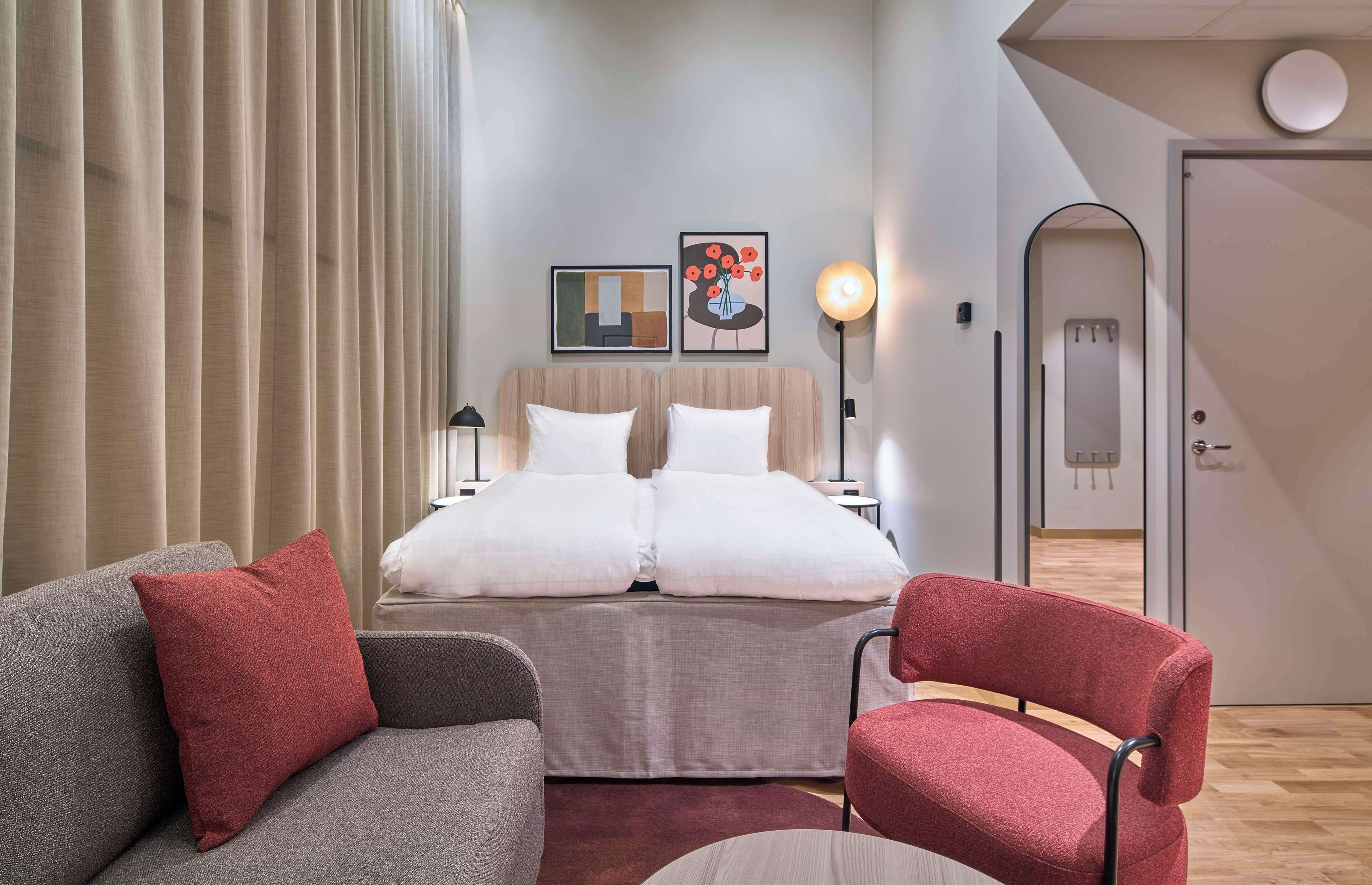 Una habitación de hotel moderna con una cama, dos mesitas de noche, un escritorio, una silla y un ventanal con cortinas.