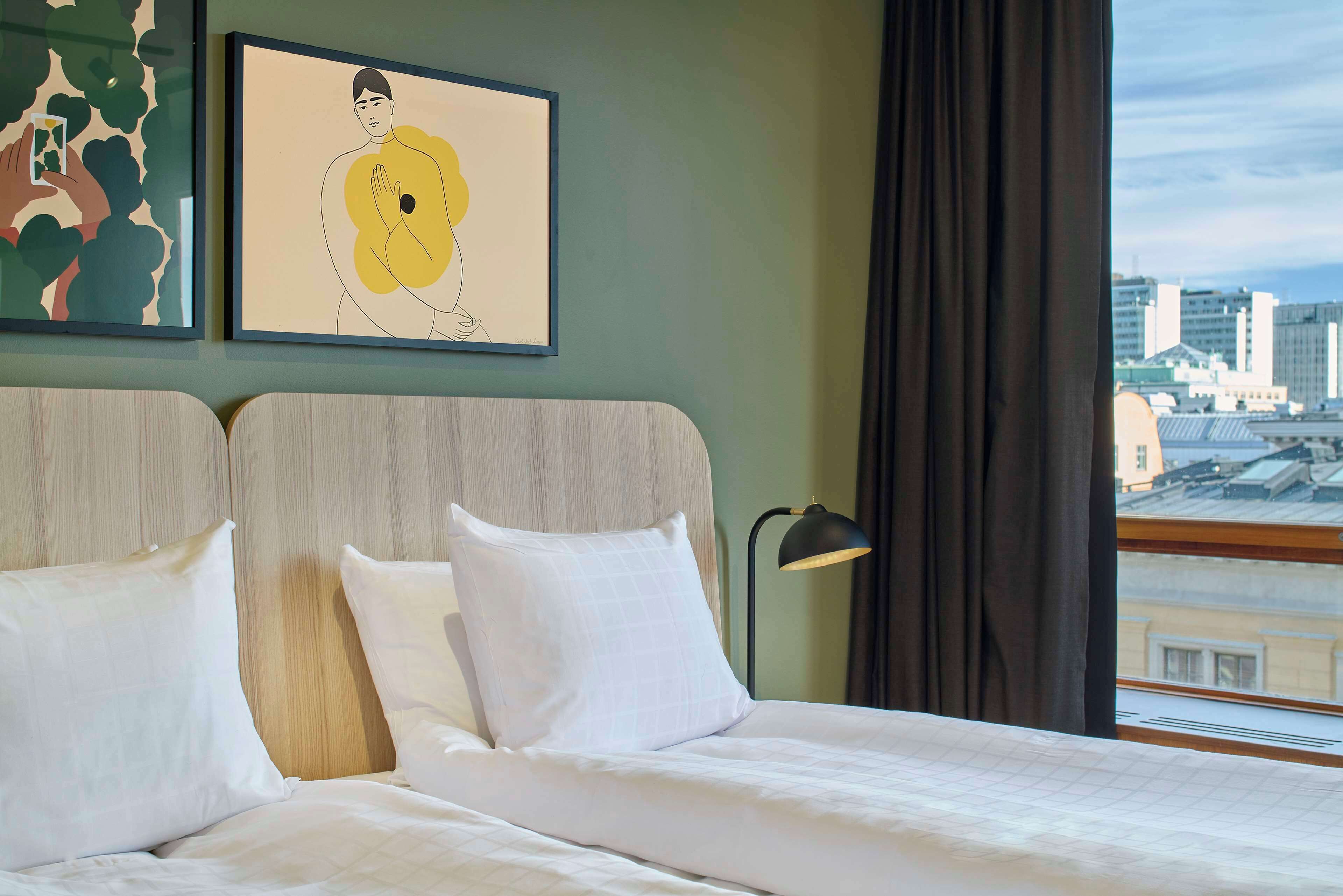 A hotel room with a bed, two pillows, a nightstand with a lamp, and two framed artworks on the wall.