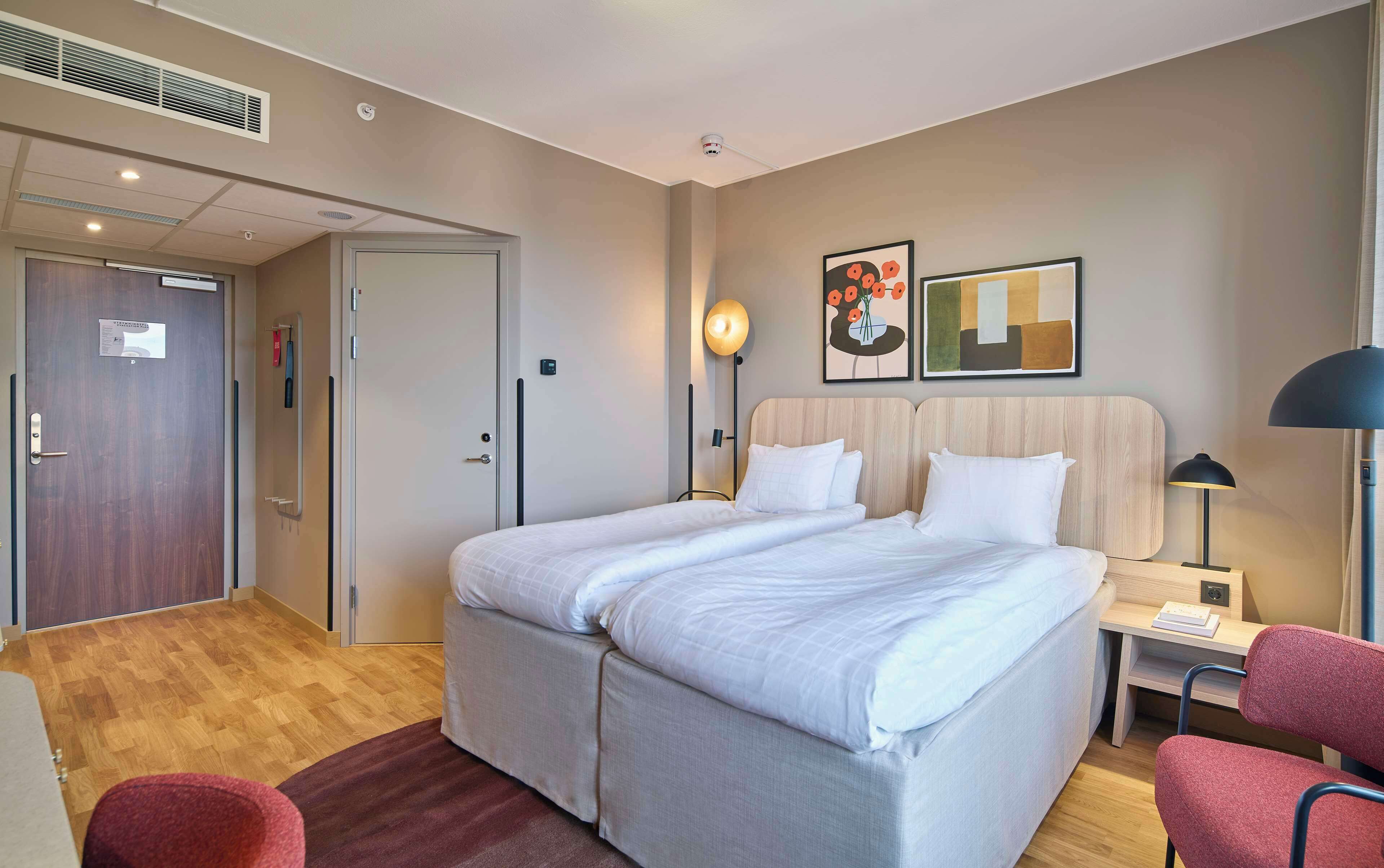 Habitación de hotel con una cama grande, un escritorio con silla, una silla roja, una lámpara de pie y obras de arte en la pared.
