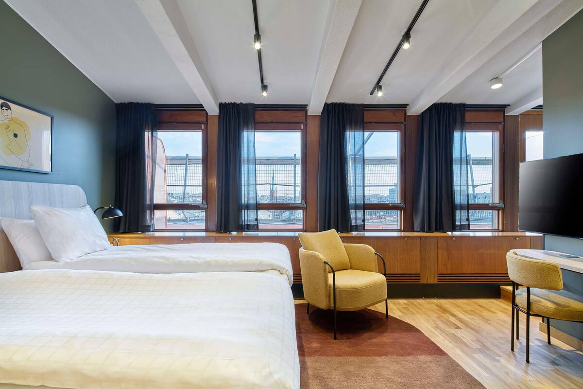 A hotel room with two beds, a chair, a TV, and large windows with a city view.