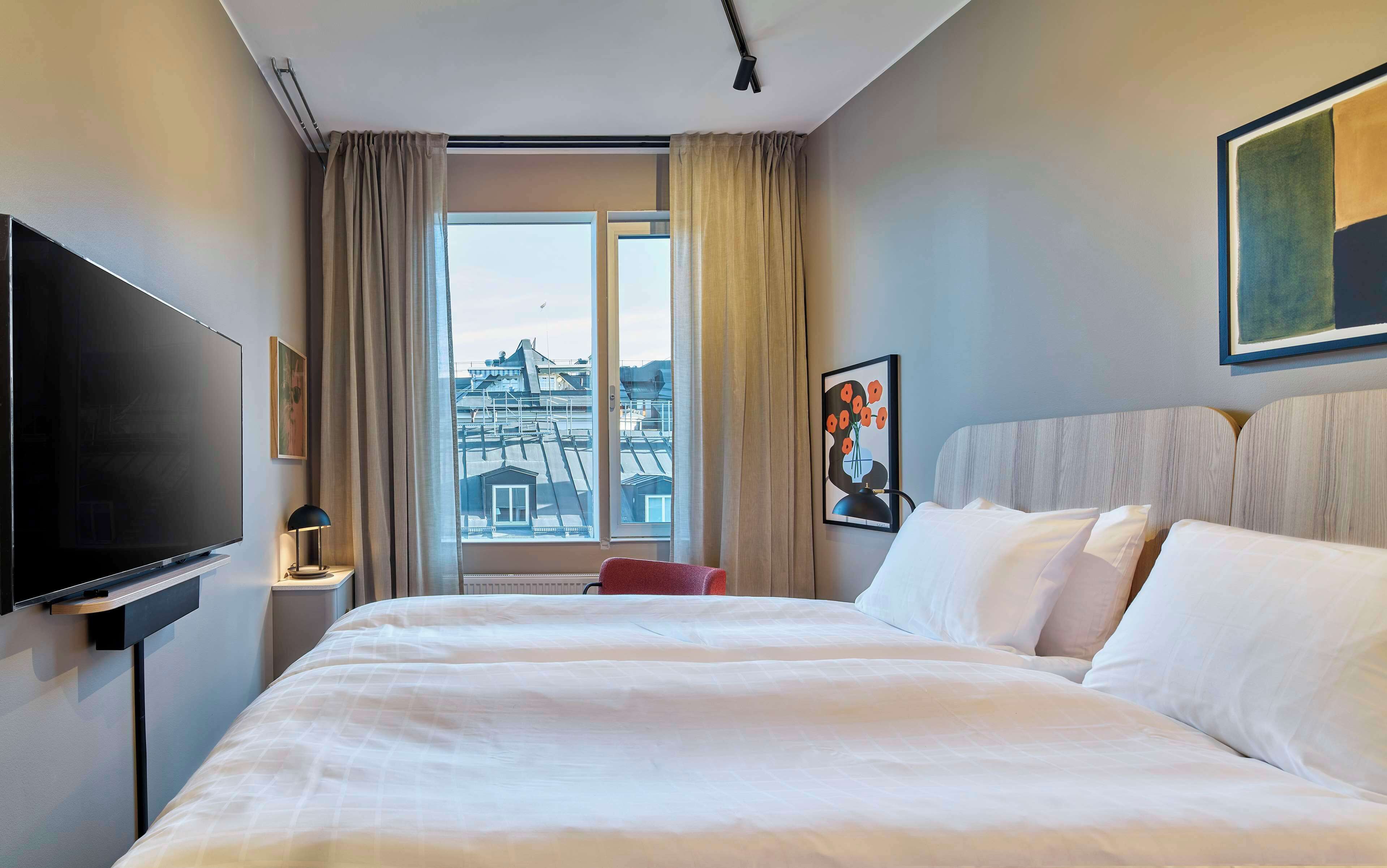 Una habitación de hotel con una cama grande, un televisor de pantalla plana, una ventana con vista a edificios y un cuadro en la pared.