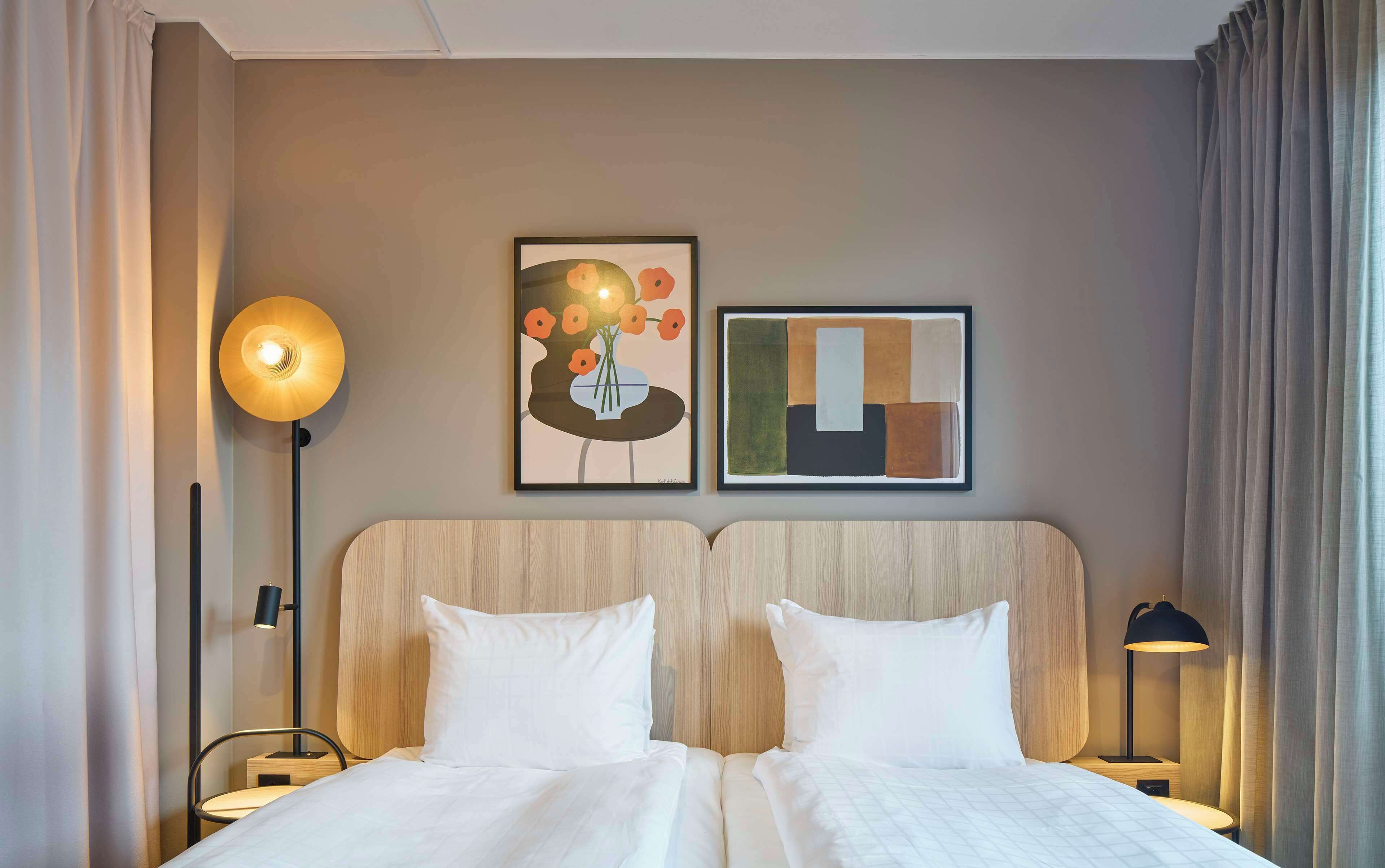 Habitación de hotel con cama doble, dos mesitas de noche, una lámpara de pie y dos cuadros en la pared.