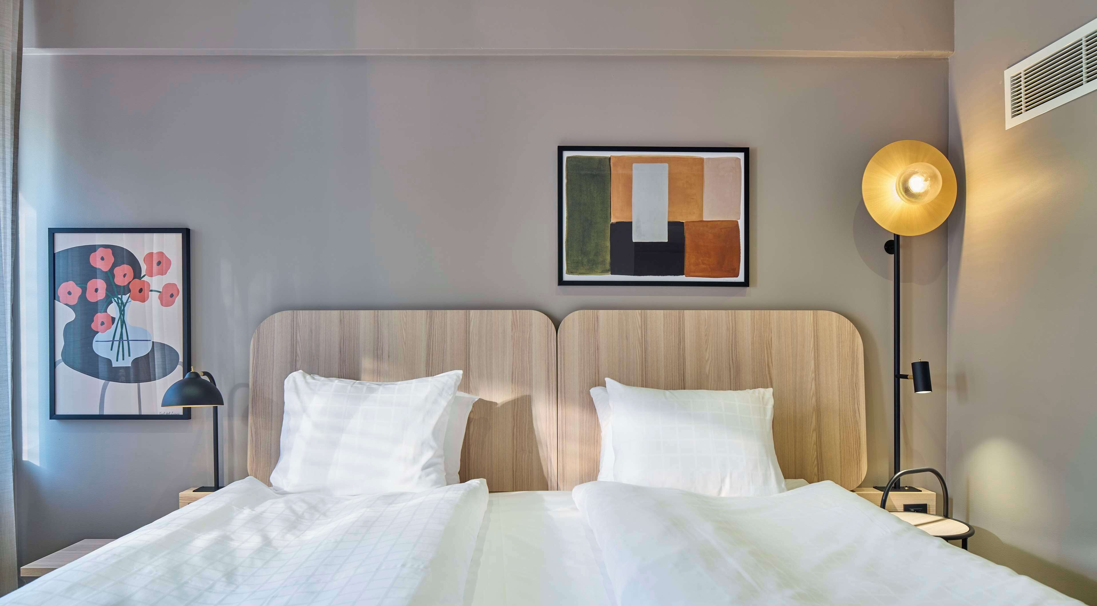 Habitación de hotel con cama, dos almohadas, un cuadro abstracto enmarcado, una lámpara de pie y una luz empotrada en la pared.