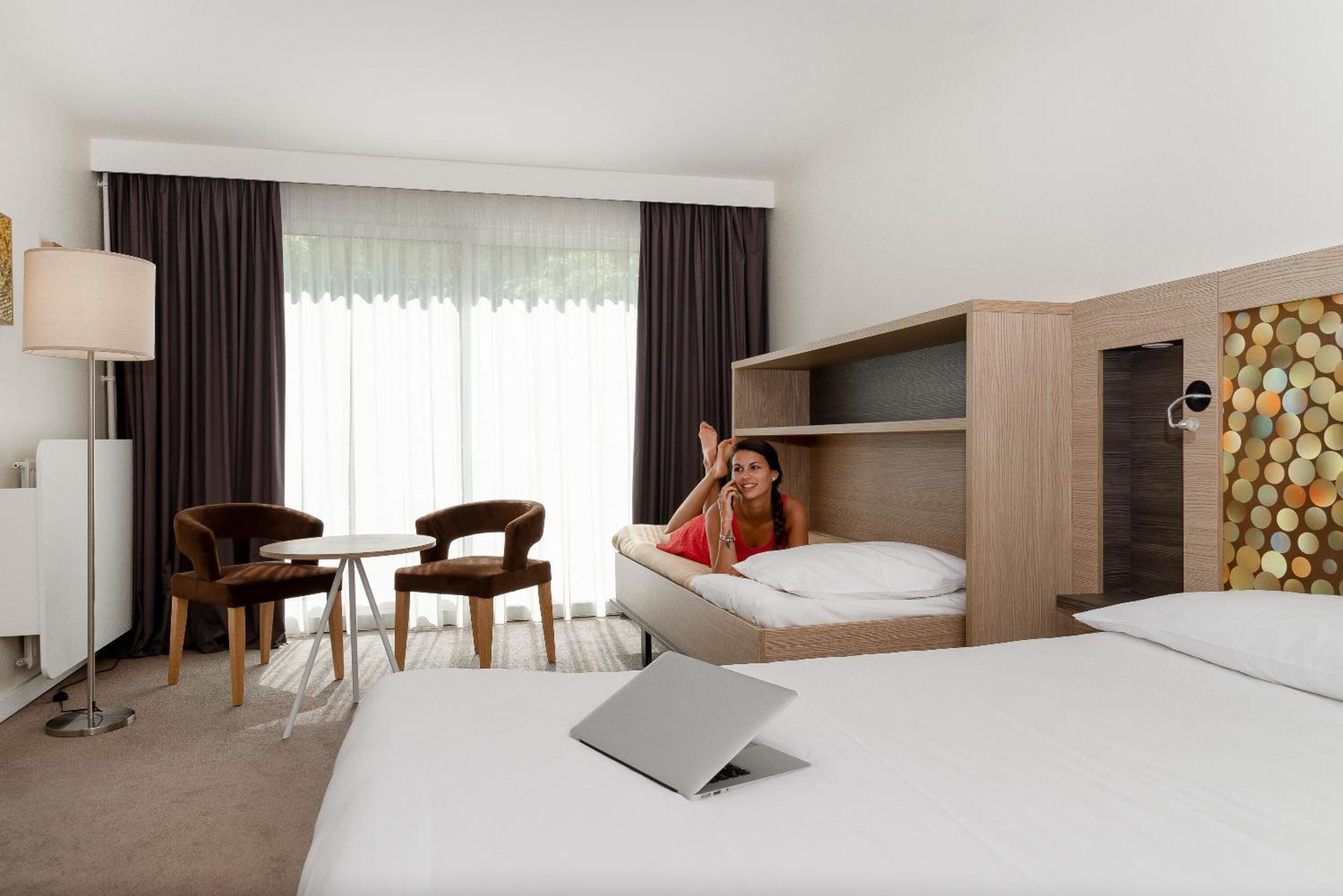 Una habitación de hotel con una cama, un escritorio, una silla, una computadora portátil y una persona sentada en la cama.