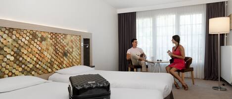 A hotel room with two beds, a large wall art piece, a suitcase on one bed, and a table with two people.