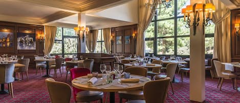 A spacious dining area with multiple tables and chairs, large windows with curtains, and wall-mounted lighting fixtures.