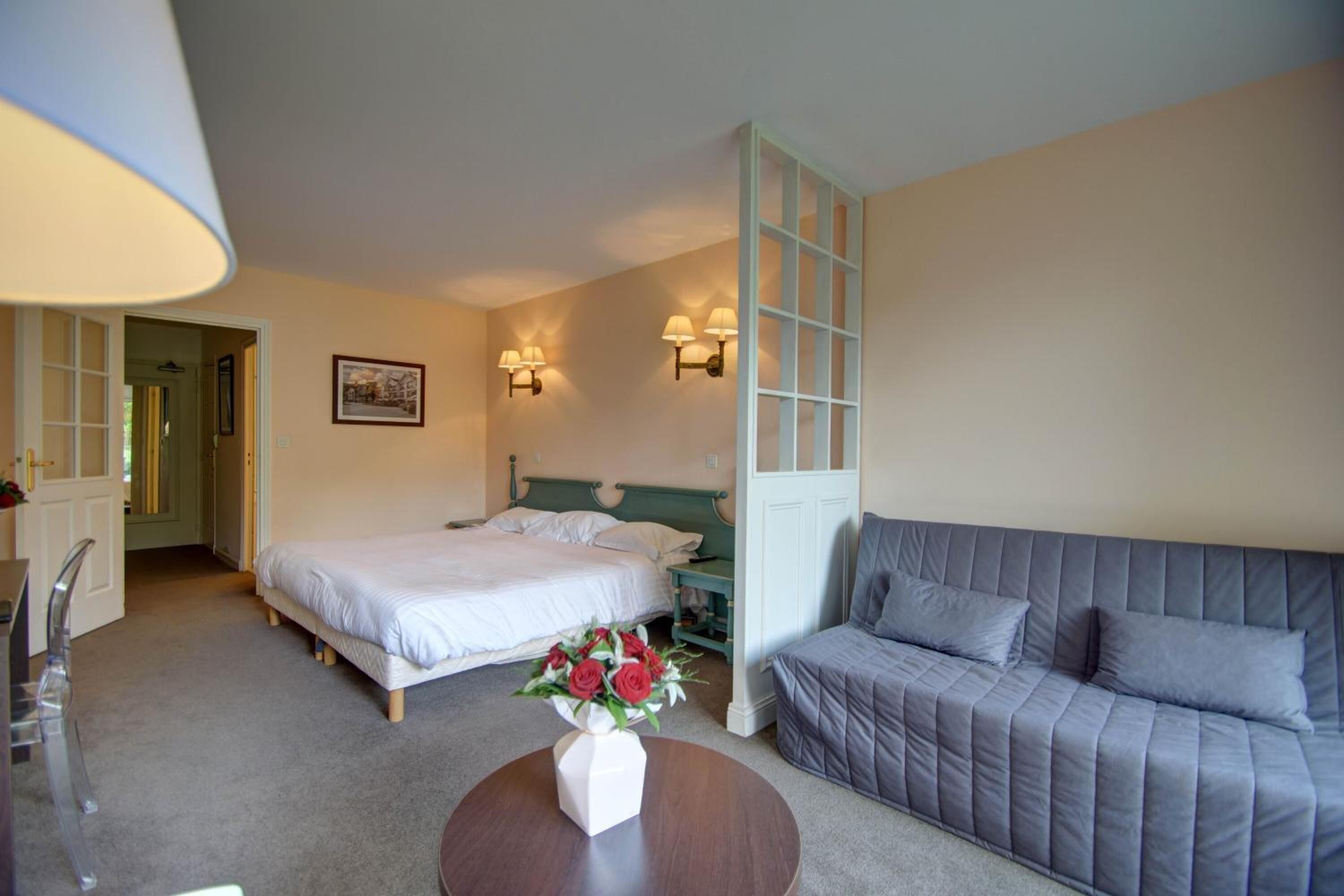A hotel room with a bed, a grey sofa, a small table with a vase of flowers, and two wall-mounted lamps.