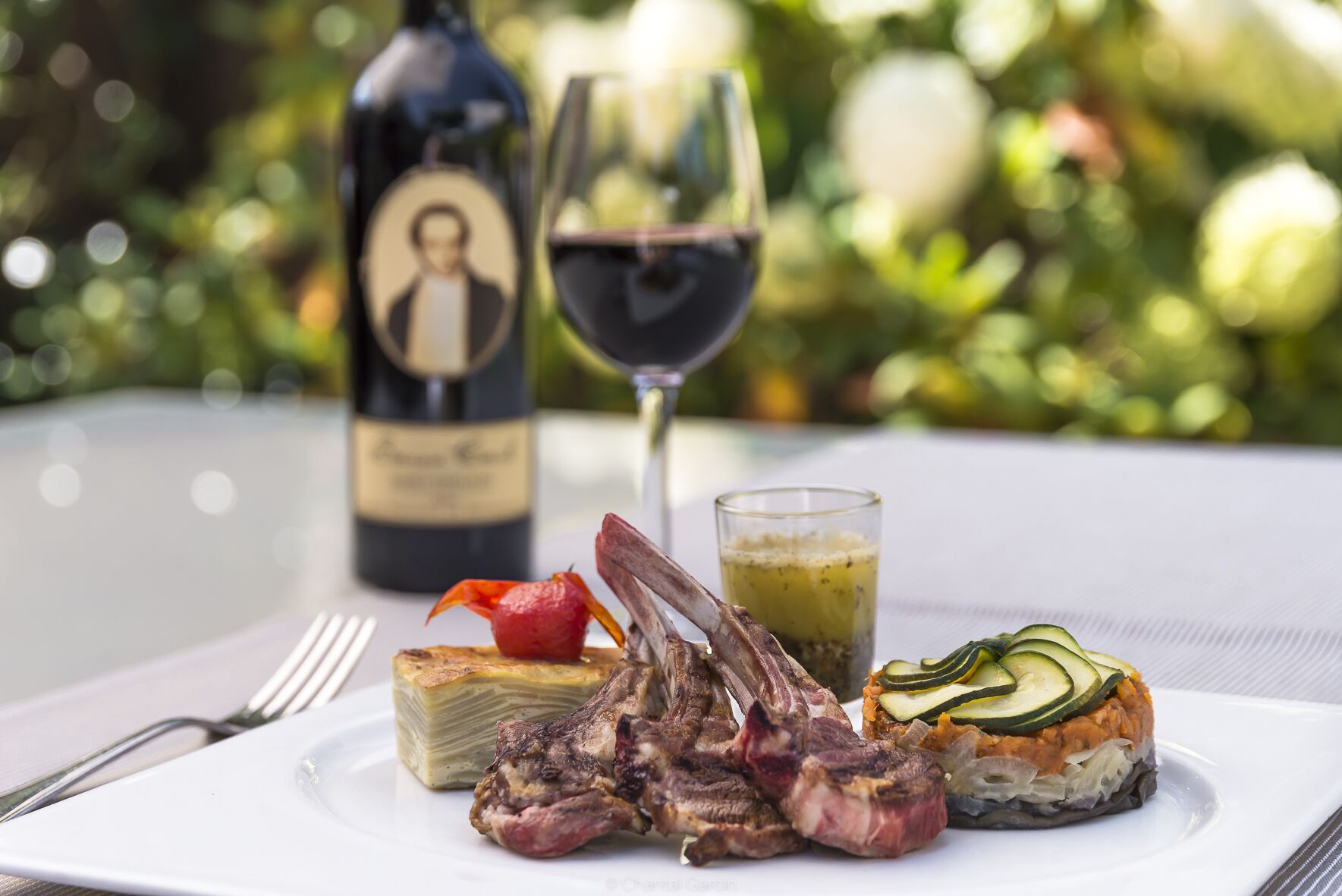 A plate with lamb chops, a stuffed vegetable, and a glass of red wine on a table with a blurred garden background.
