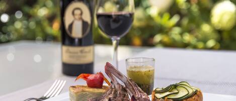 A plate with lamb chops, a stuffed vegetable, and a glass of red wine on a table with a blurred garden background.