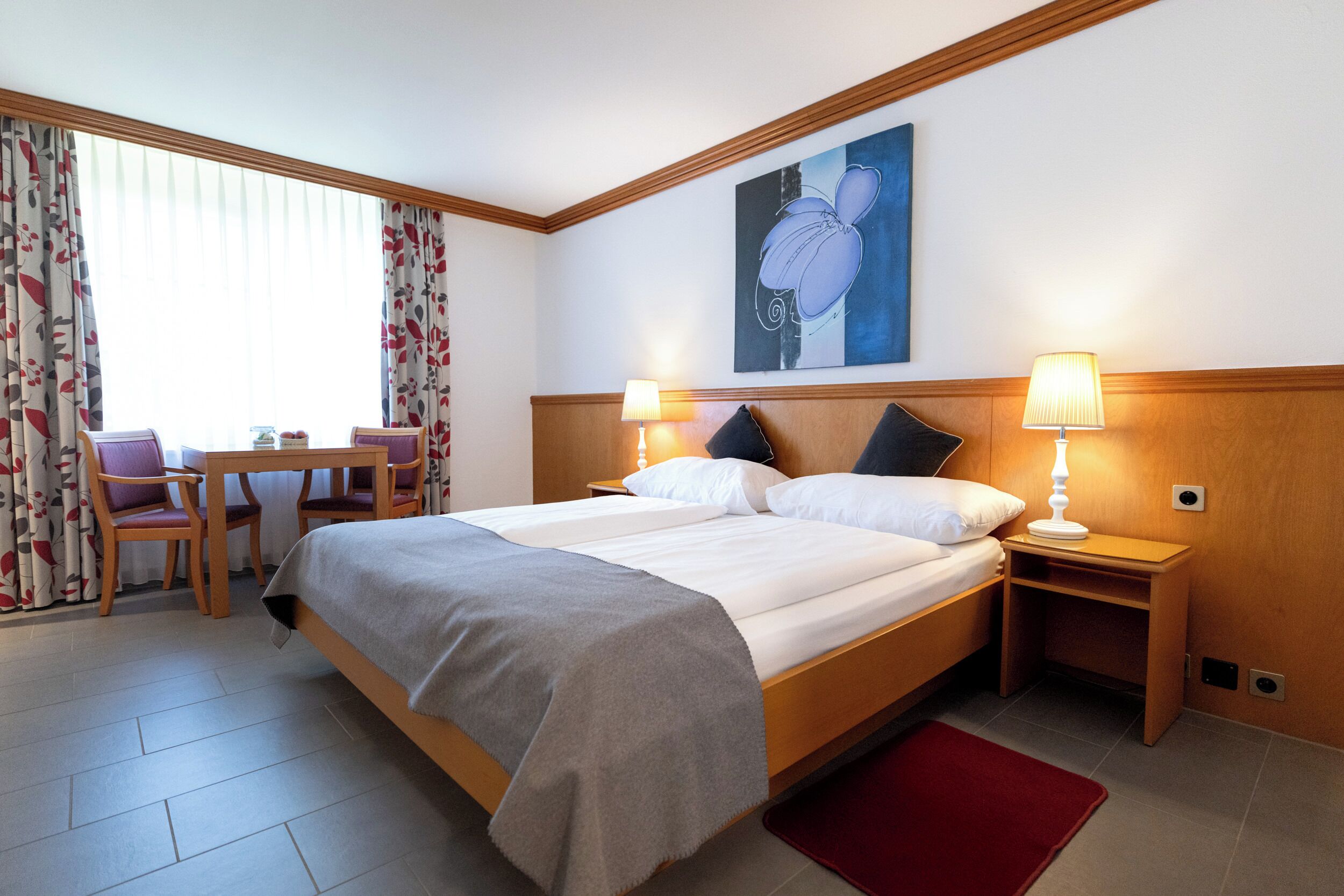 A hotel room with a large bed, two bedside tables with lamps, a desk with a chair, and a window with curtains.