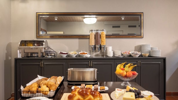 Montaje de bufé con diversas opciones de comida, incluyendo pasteles, frutas y quesos, dispuesto sobre una mesa con una estación de café y un microondas al fondo.