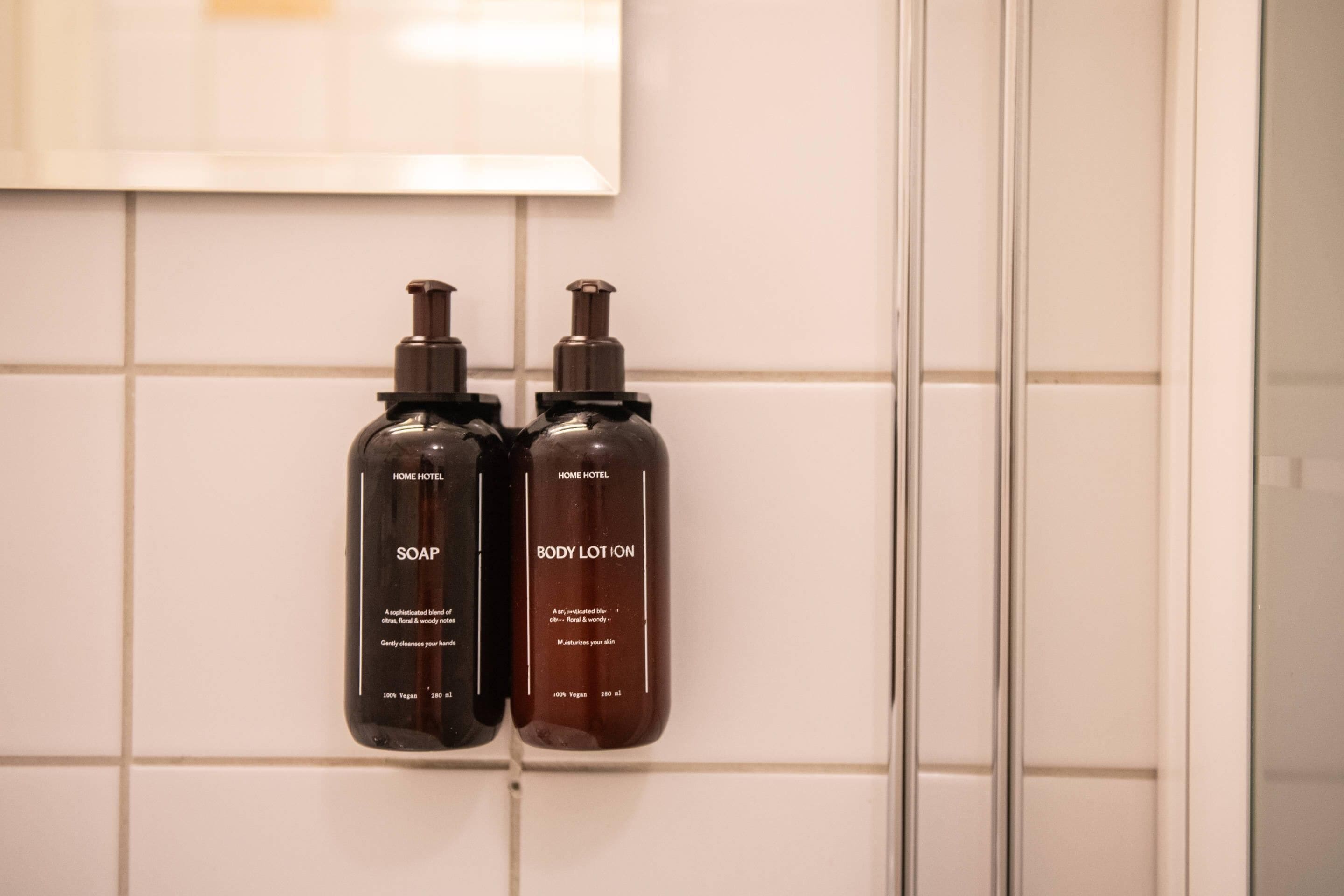 Two bottles of soap and body lotion on a tiled wall.