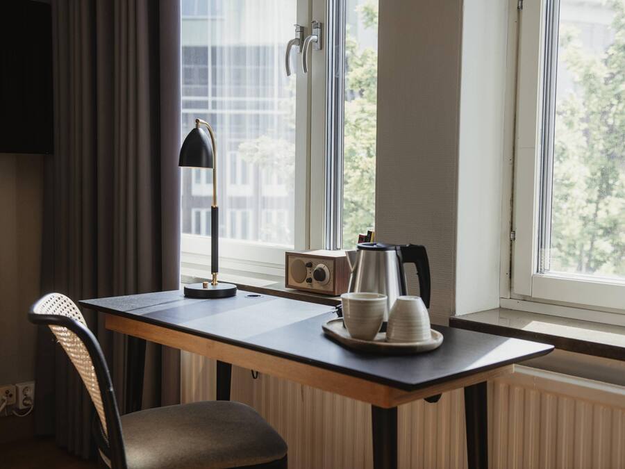 Una habitación pequeña y moderna con un escritorio de madera, una silla, una lámpara, una radio y una ventana que ofrece vistas a edificios y árboles.