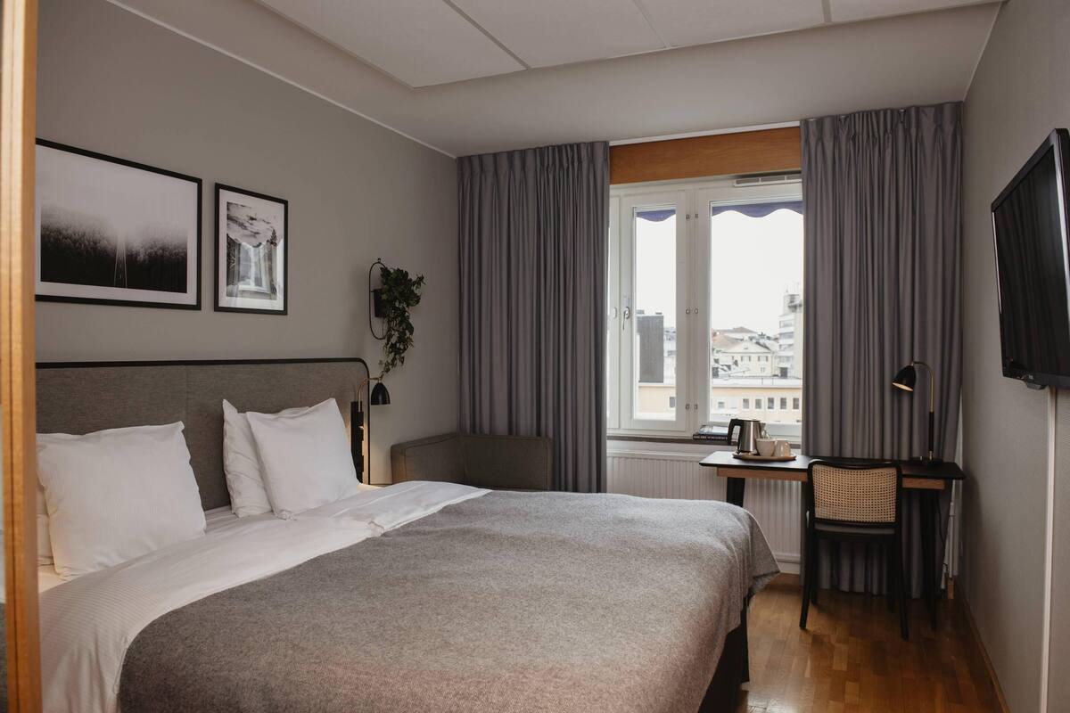 Una habitación de hotel con cama, escritorio, silla, televisor y ventana con cortinas.