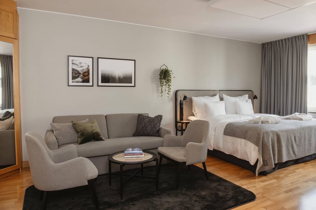 Una habitación de hotel moderna con un sofá gris, una pequeña mesa redonda con libros, una cama con ropa de cama blanca y dos cuadros en la pared.