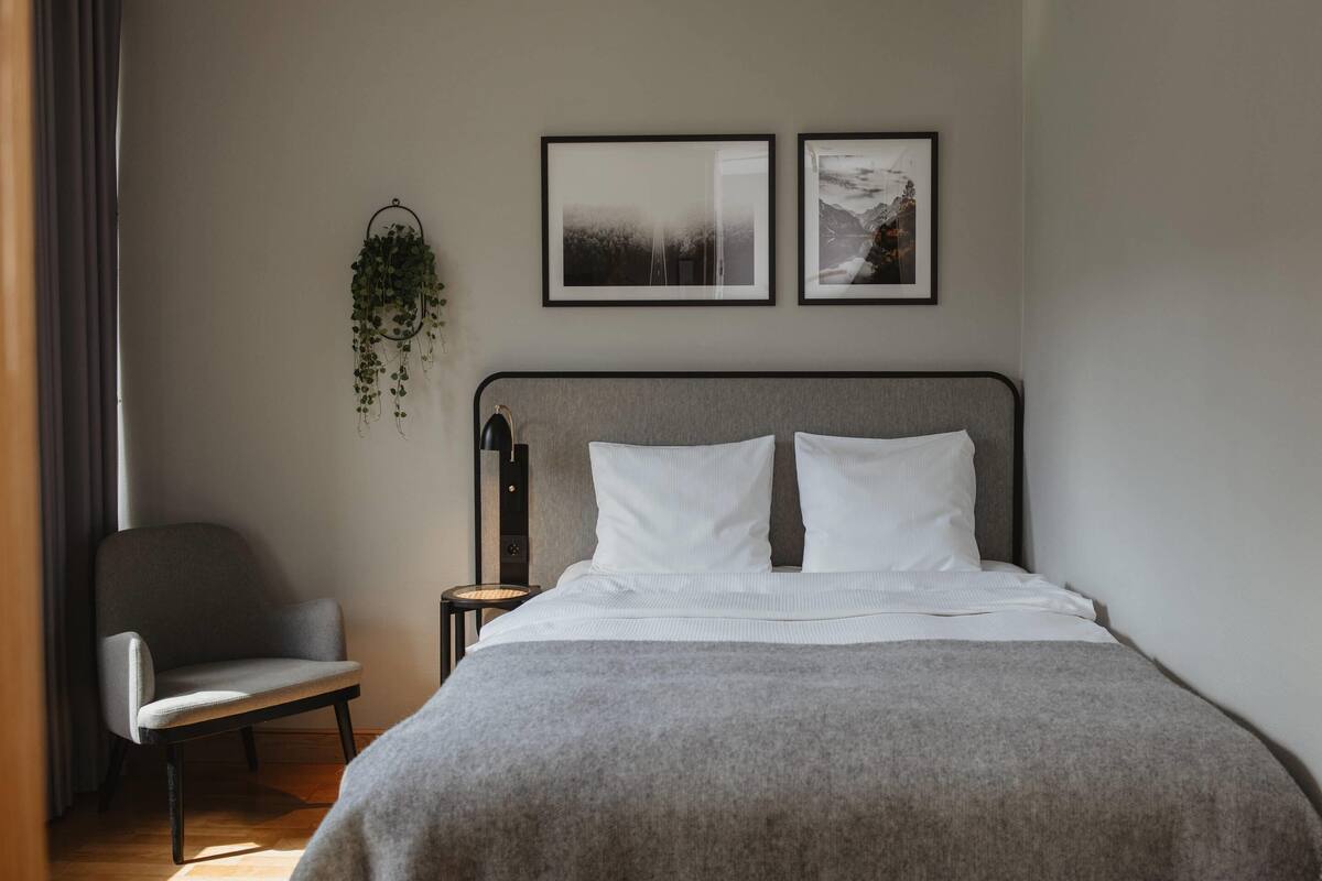 Un dormitorio con una cama, una silla, una mesita de noche y dos cuadros enmarcados en la pared.