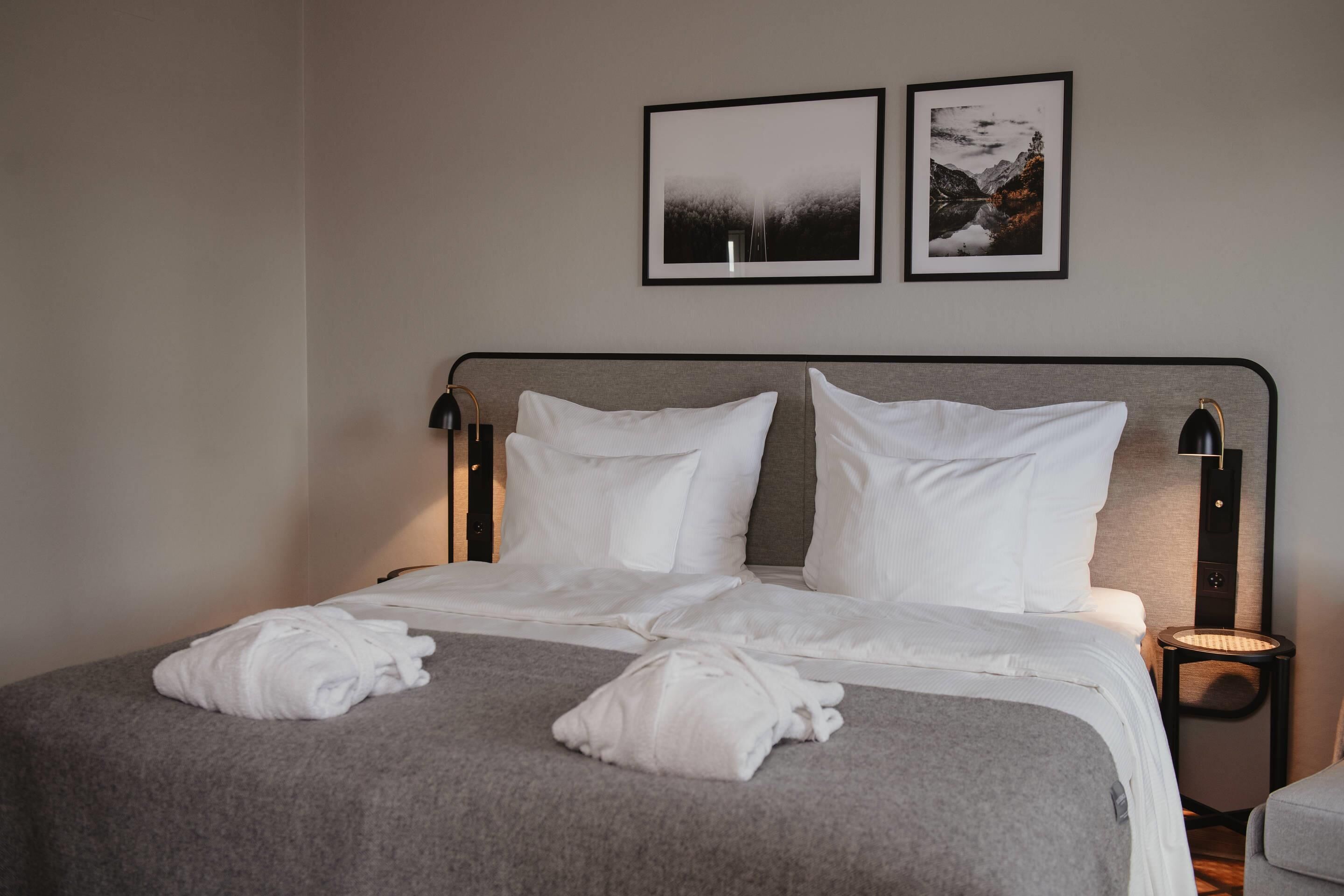 Una cama bien hecha con sábanas blancas y dos almohadas, un plaid gris y dos toallas enrolladas sobre la cama. Dos cuadros enmarcados están colgados en la pared sobre la cama.