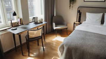 A hotel room with a bed, a desk with a chair, a small table with a radio, a window with curtains, and a plant on the wall.