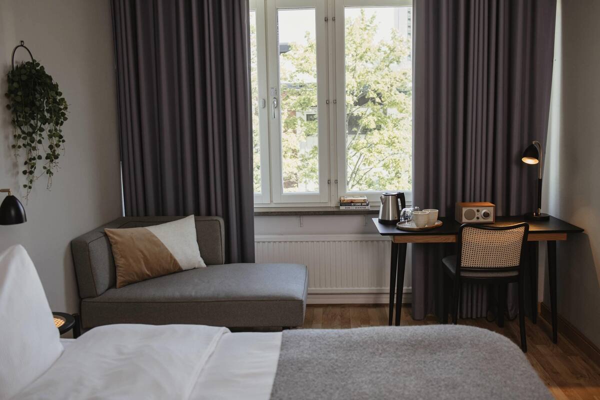 Un dormitorio con cama, sofá, escritorio con silla, radiador, ventana con cortinas y una planta colgante.