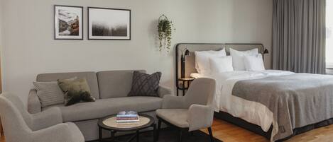 Un dormitorio moderno con un sofá gris, una mesita redonda pequeña, una cama con ropa blanca y dos cuadros enmarcados en la pared.