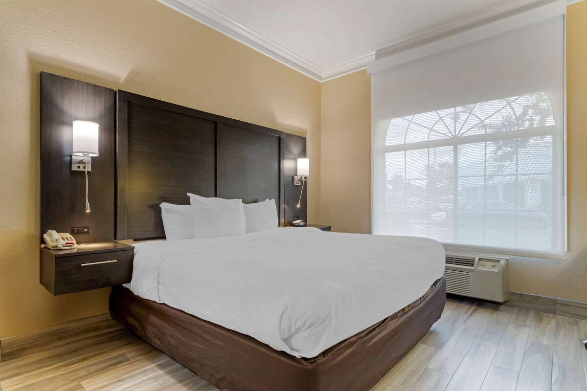 Ein Hotelzimmer mit einem großen Bett, einem dunklen Holz-Kopfschild, Nachttischlampen, einem Telefon und einem Fenster mit Jalousien.