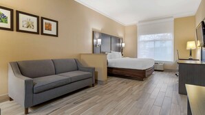 Una habitación de hotel con un sofá gris, una cama con cabecera de madera, un escritorio con lámpara y cuadros enmarcados en la pared.