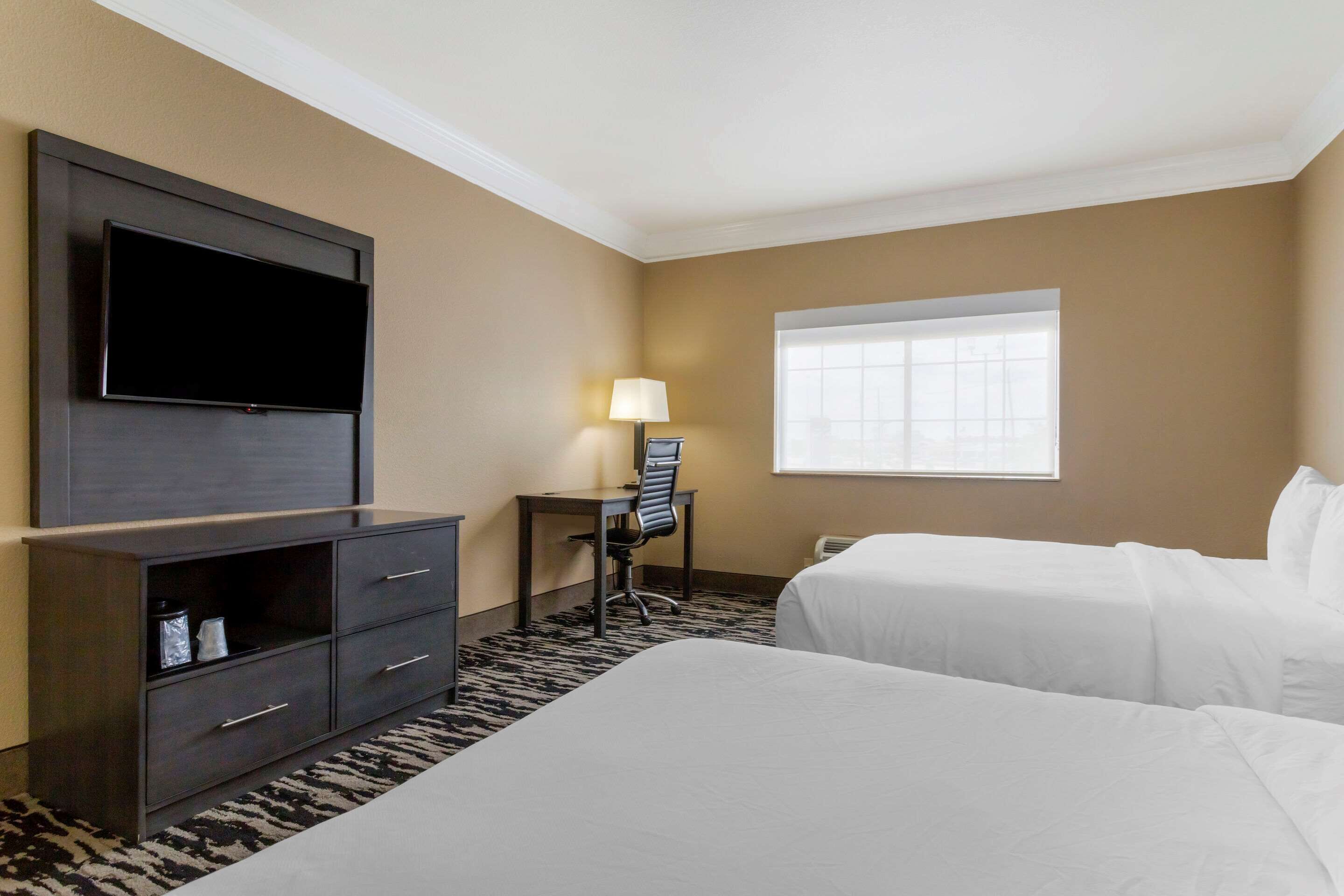 Une chambre d’hôtel dotée d’un grand téléviseur à écran plat fixé au mur, d’un bureau avec une chaise, de deux lits, d’une fenêtre avec des stores et d’un tapis à motifs.