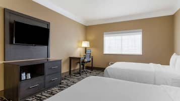 Une chambre d’hôtel dotée d’un grand téléviseur à écran plat fixé au mur, d’un bureau avec une chaise, de deux lits, d’une fenêtre avec des stores et d’un tapis à motifs.
