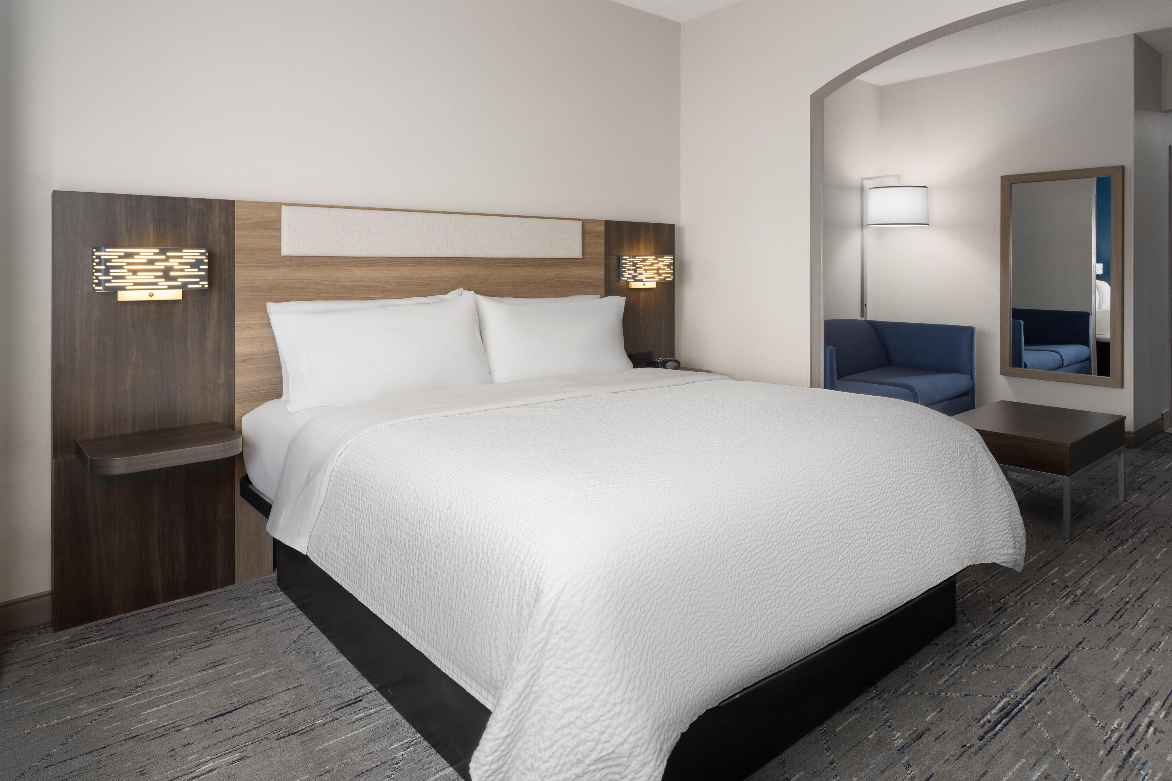 A hotel room with a large bed, a nightstand, a lamp, a mirror, and a blue chair.