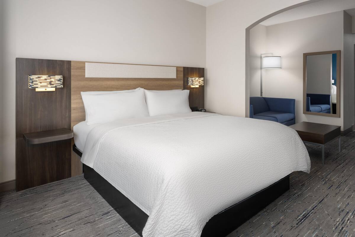 Une chambre d’hôtel avec un grand lit, une table de chevet, une lampe, un miroir et une chaise bleue.