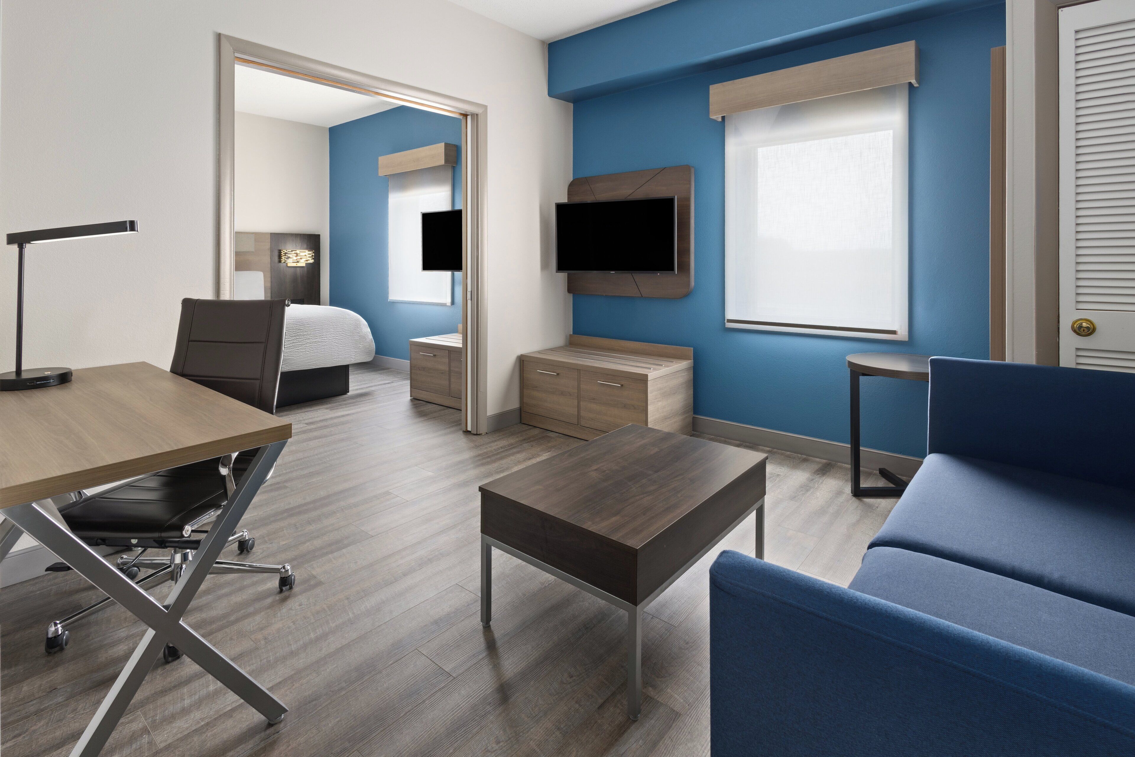 A modern hotel room with a wooden desk, a blue sofa, a bed, a TV mounted on the wall, and a small table.