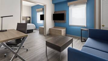 A modern hotel room with a wooden desk, a blue sofa, a bed, a TV mounted on the wall, and a small table.