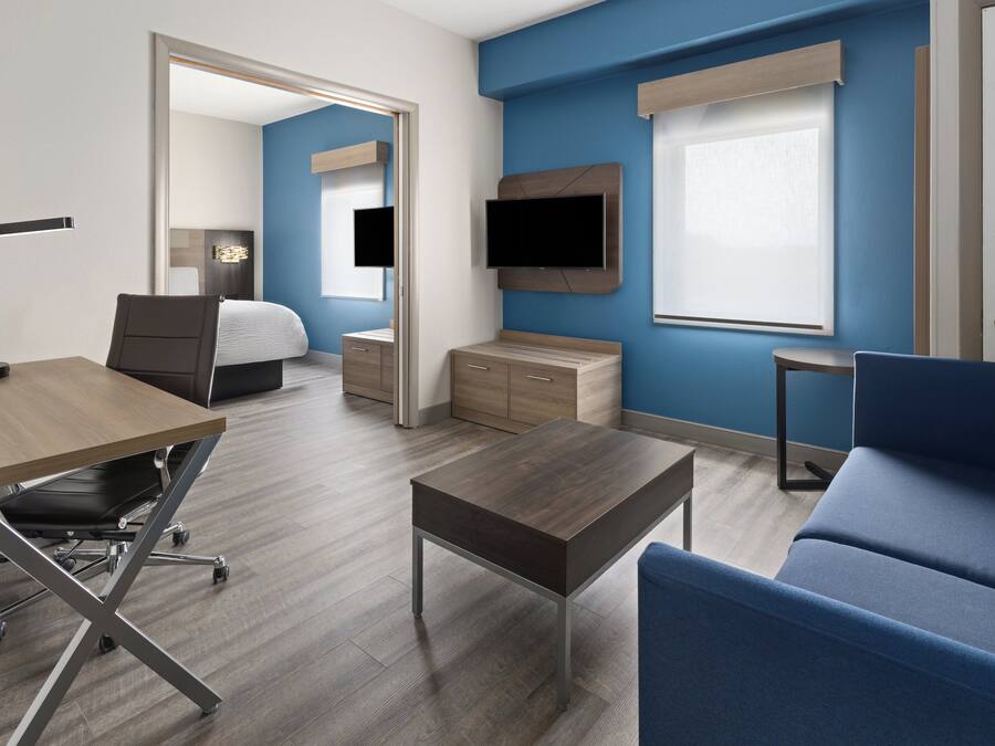 A modern hotel room with a wooden desk, a blue sofa, a bed, a TV mounted on the wall, and a small table.
