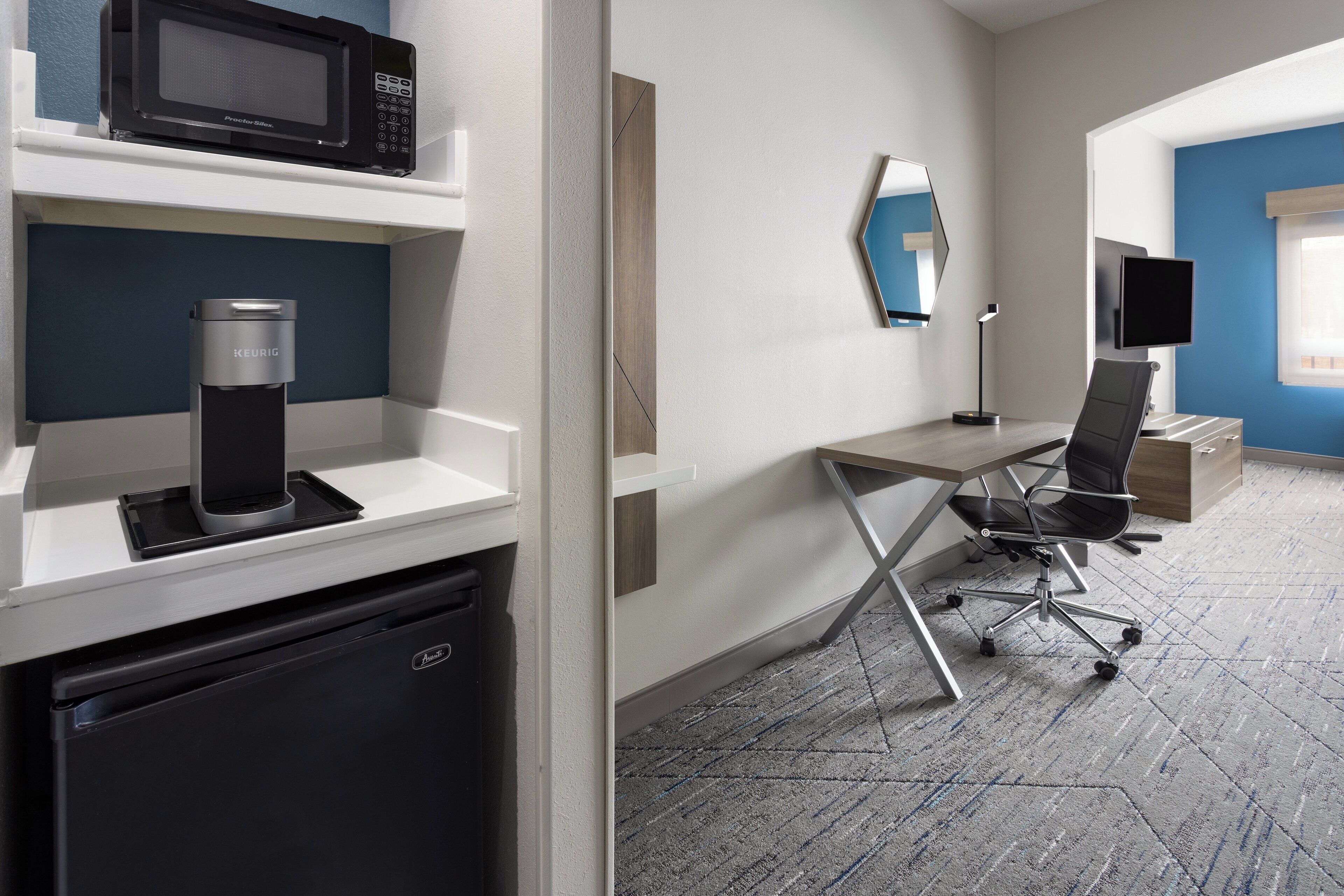 A hotel room with a kitchenette, a microwave, a Keurig coffee maker, a desk with a chair, a flat-screen TV, and a large mirror.