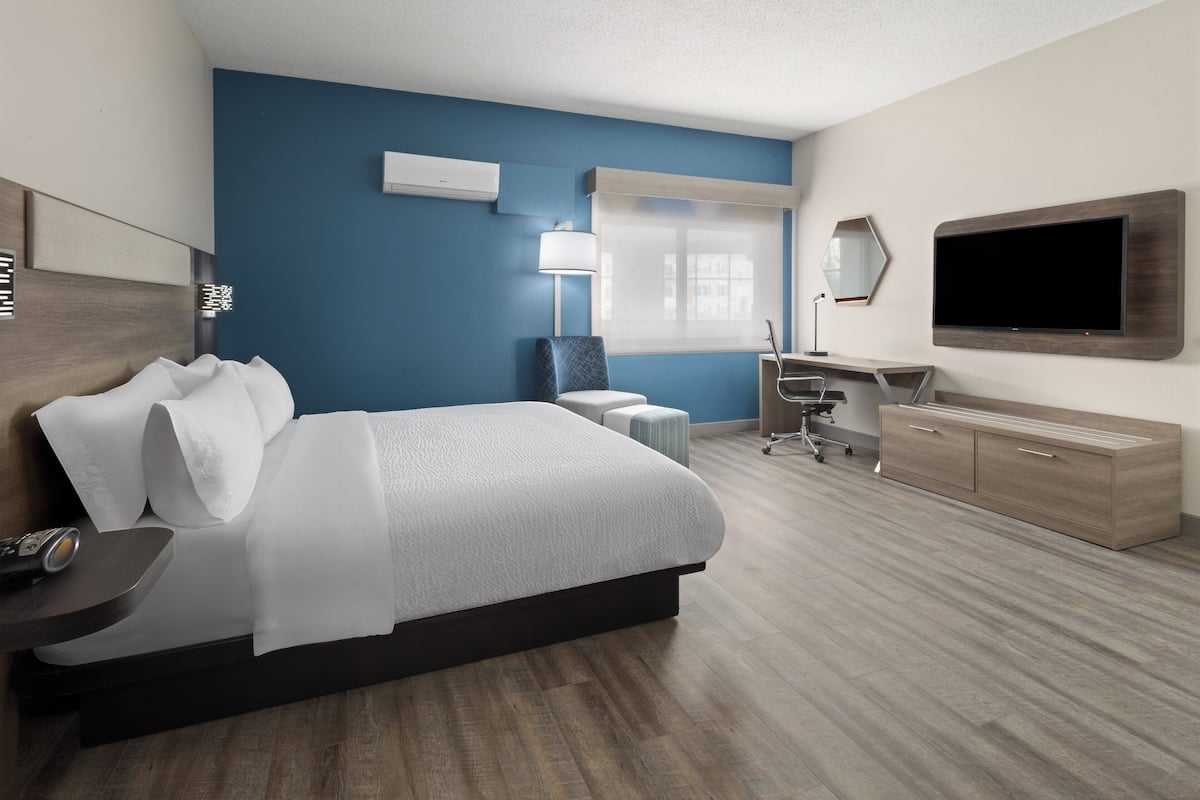 A modern hotel room with a large bed, a desk, a chair, a TV, and a window with blinds.
