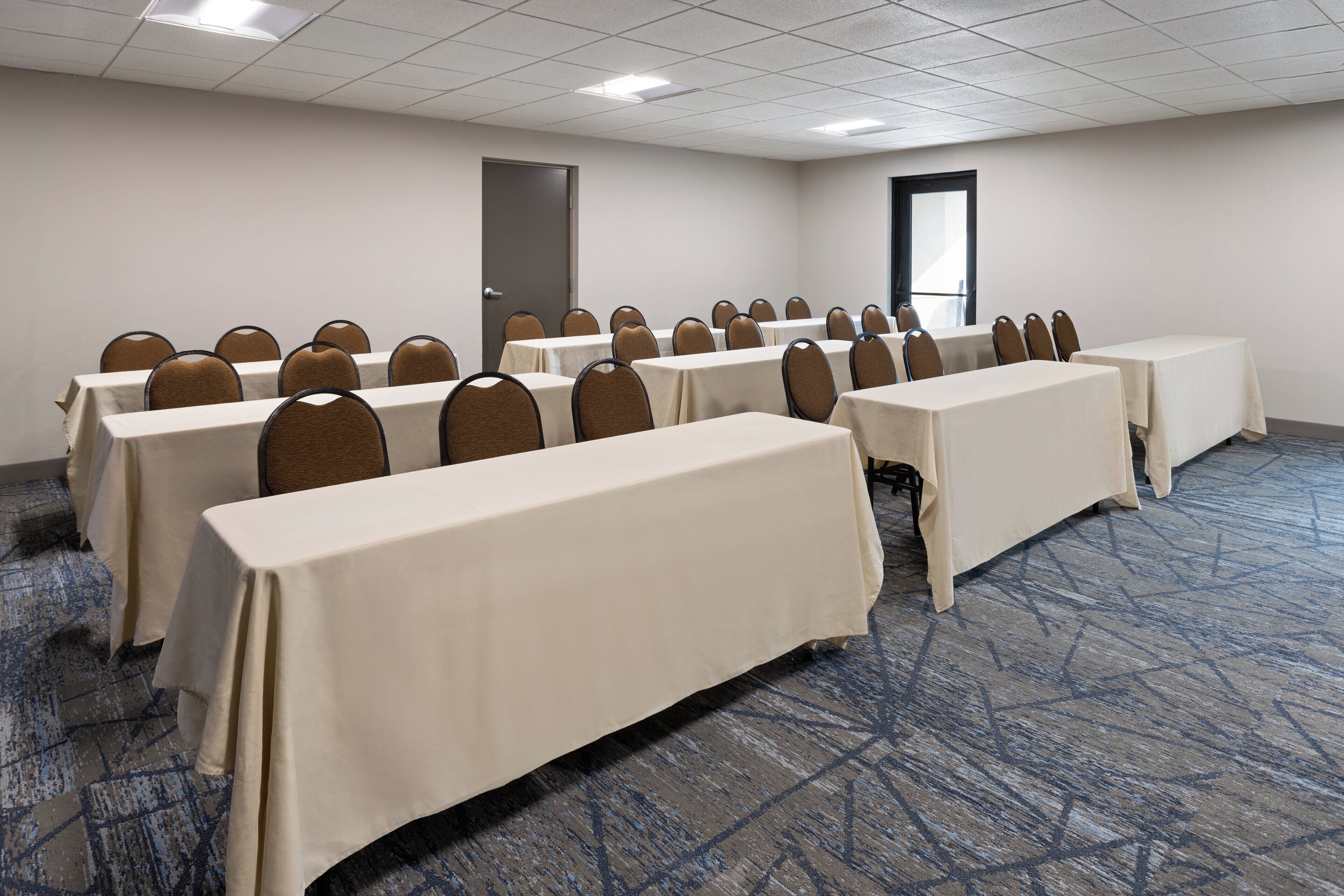 Un salón de conferencias con varias mesas y sillas, piso alfombrado y una puerta al fondo.