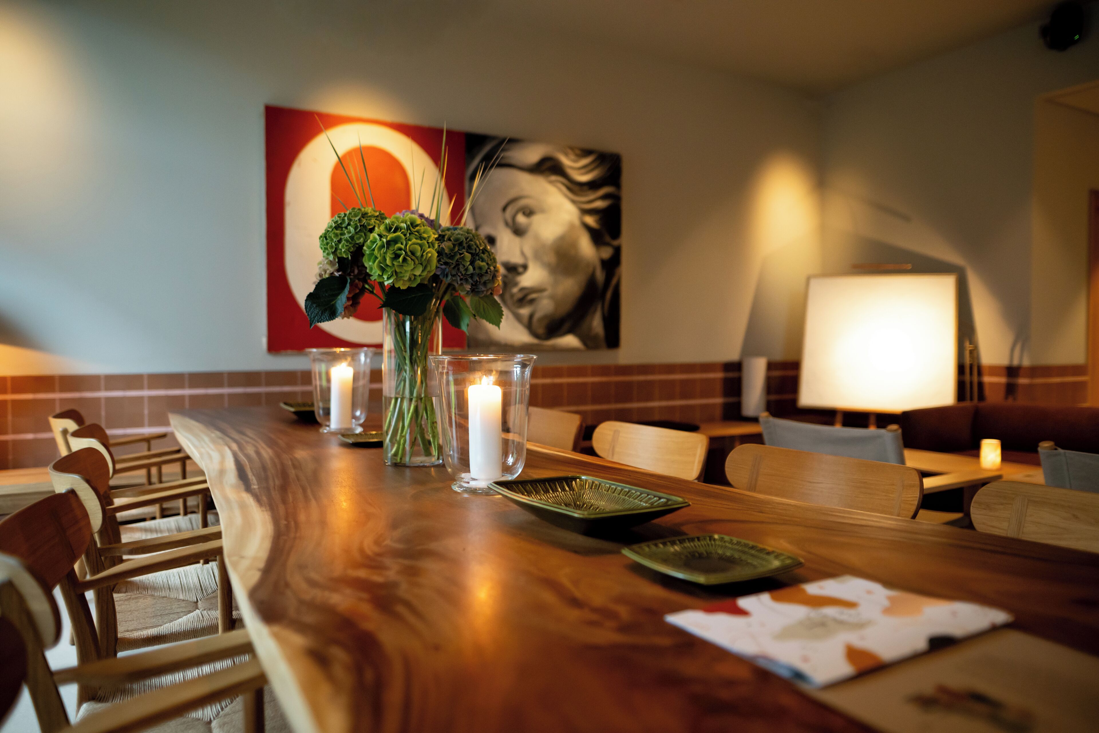 A dining area with a wooden table, chairs, a vase with flowers, a lit candle, and a painting on the wall.