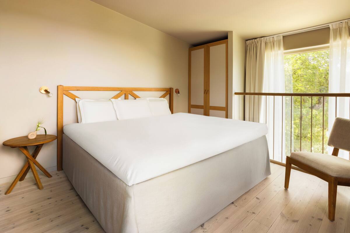 Un lit bien fait dans une pièce au plancher en bois, avec une petite table de chevet en bois, une chaise et une grande fenêtre avec des rideaux.