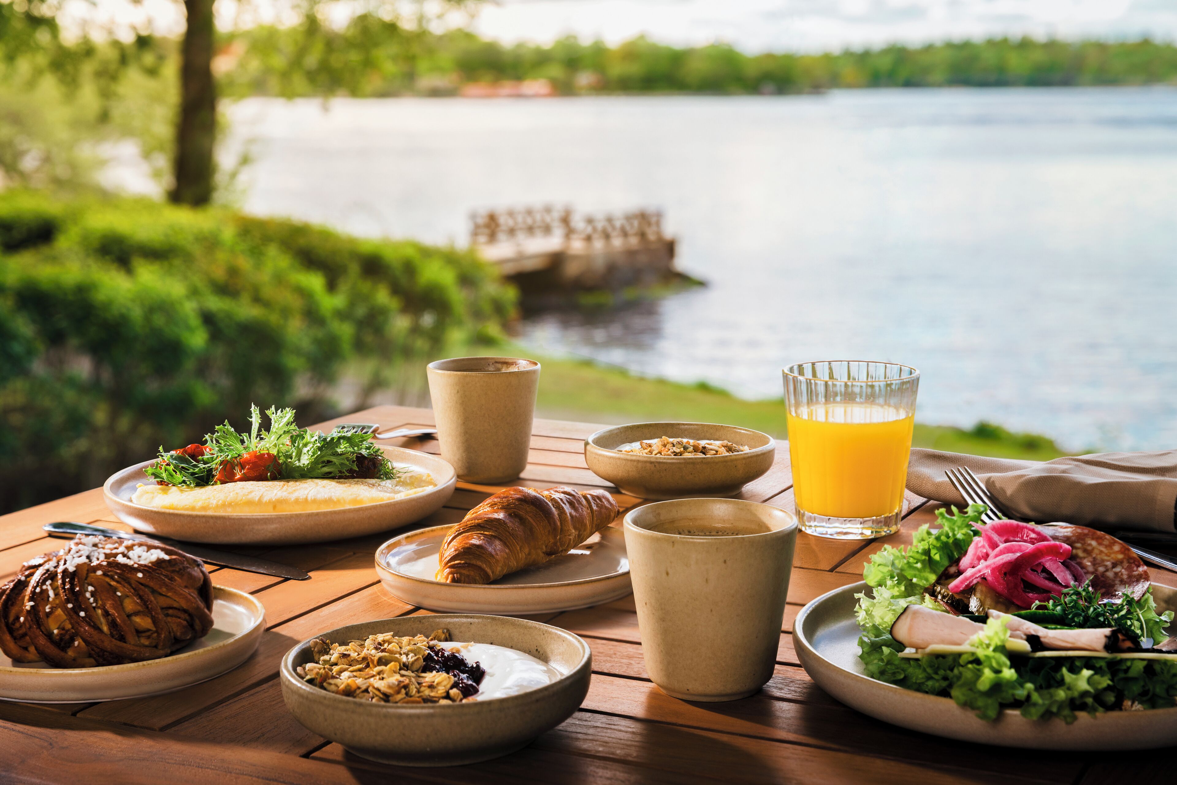 Et morgenbord med tallerkener med mad, en croissant, et glas appelsinjuice og udsigt til en sø.