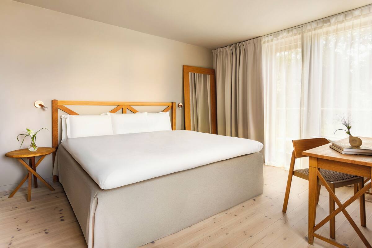 Une chambre d’hôtel avec un grand lit, une tête de lit en bois, une petite table avec un vase, une chaise et une fenêtre avec des rideaux.