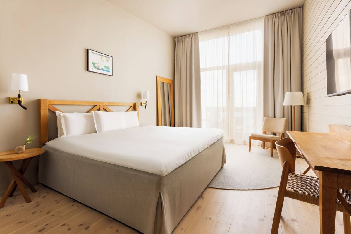 Une chambre d’hôtel avec un grand lit, un bureau en bois, une chaise, une petite table avec une lampe et un tableau au mur.