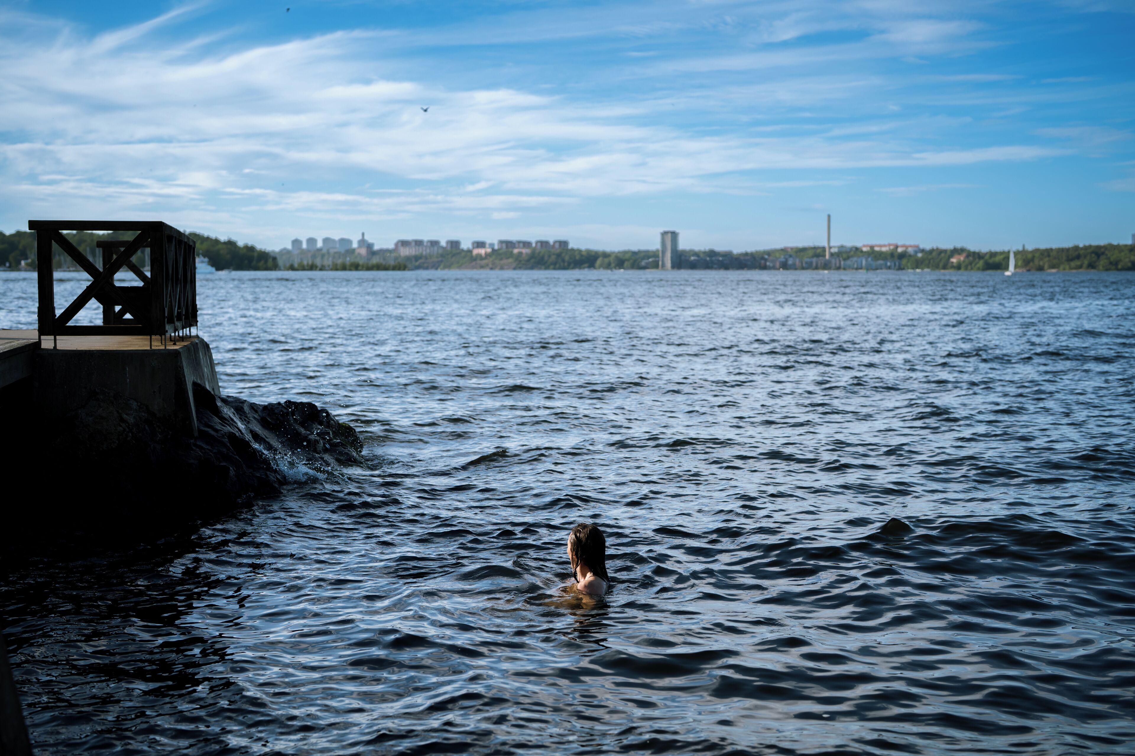 Une personne nage dans une étendue d’eau, avec un quai et des bâtiments en arrière-plan.