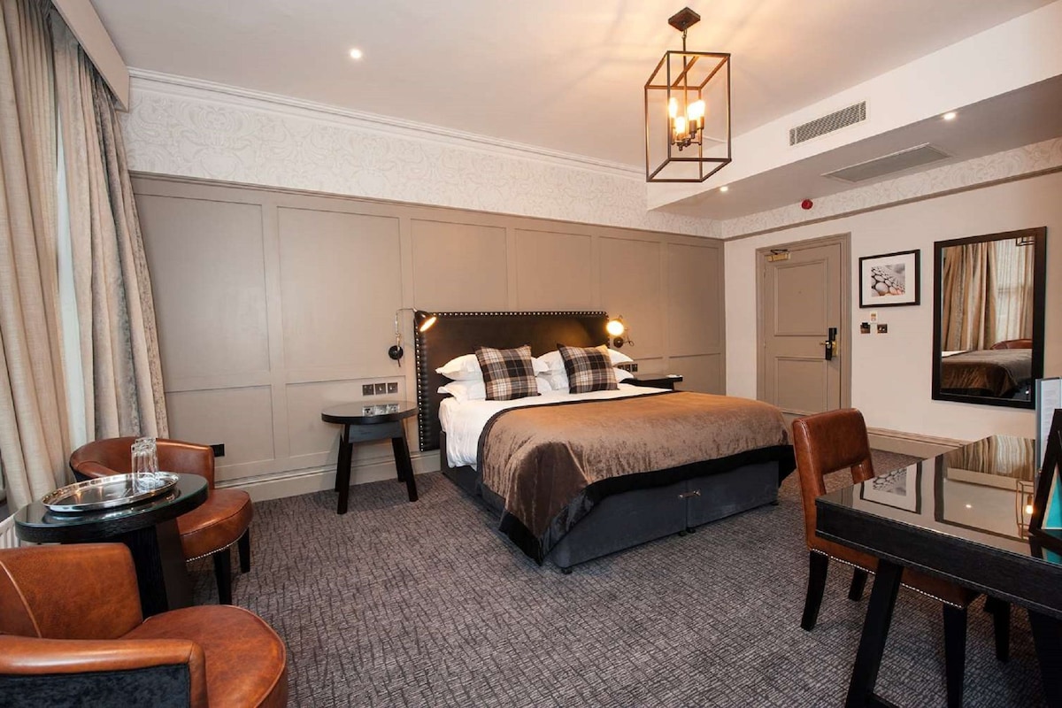 Ein Hotelzimmer mit einem großen Bett, einem Schreibtisch mit Stuhl, einem kleinen Tisch ebenfalls mit Stuhl, einem Nachttisch und einer Pendelleuchte.