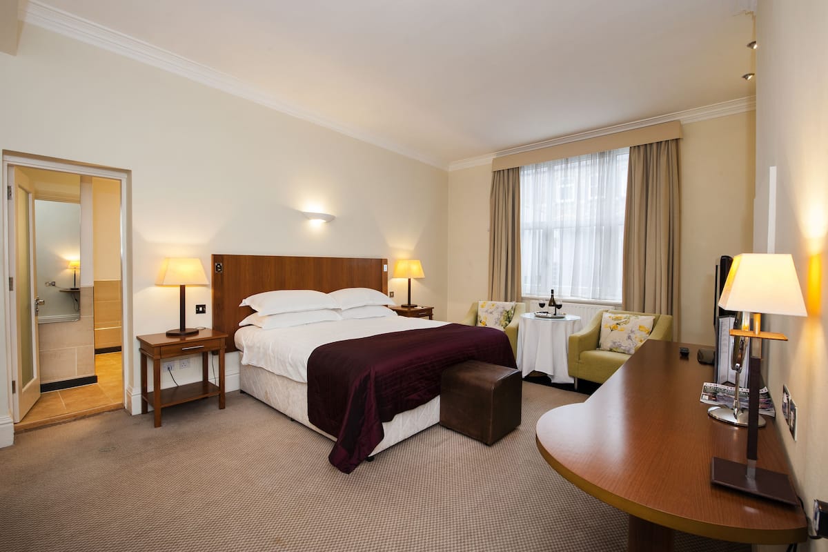 Une chambre d’hôtel avec un grand lit, deux tables de chevet avec des lampes, une petite table ronde avec une chaise, une fenêtre avec des rideaux et une porte donnant sur une salle de bain.