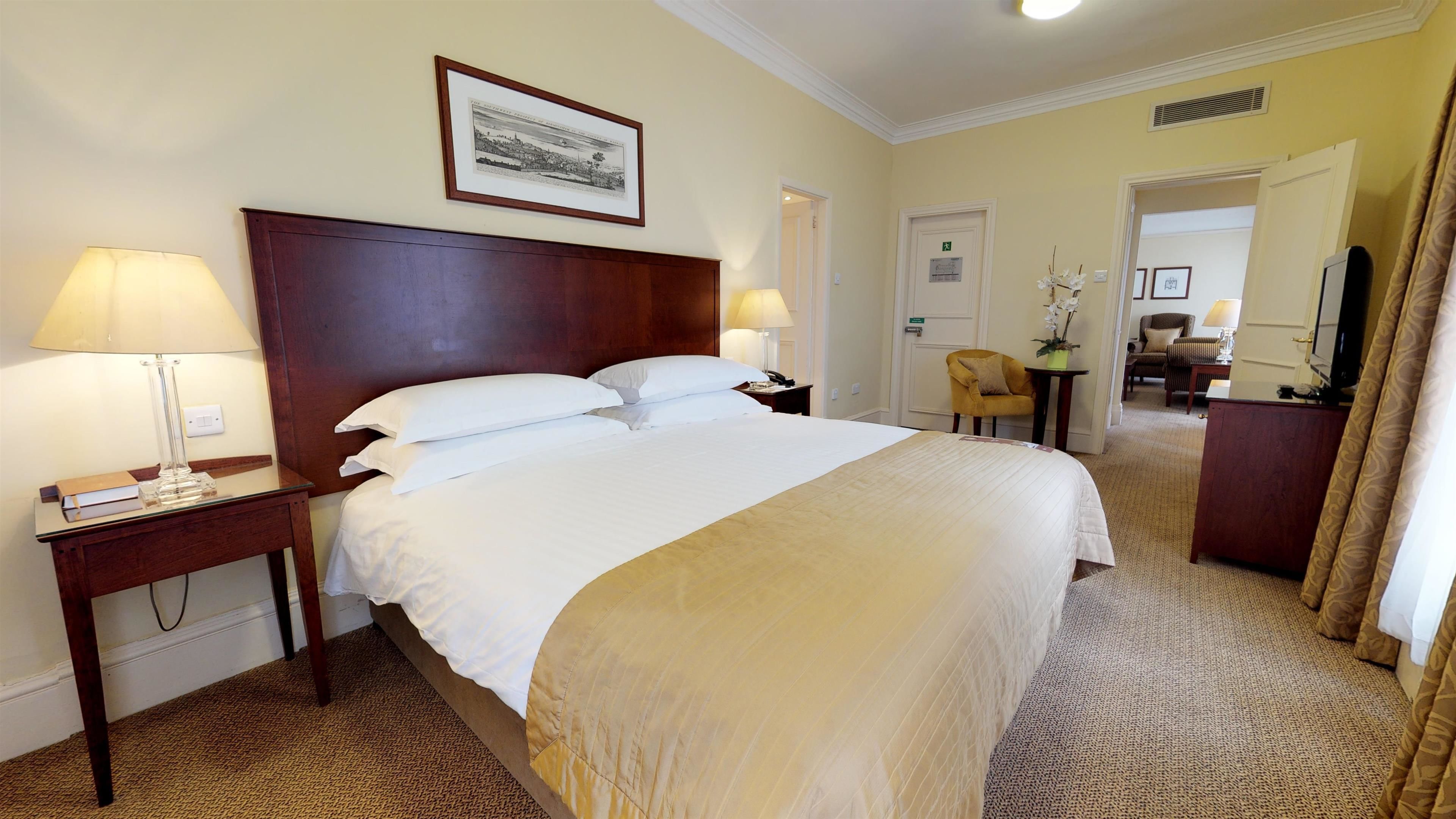 A hotel room with a large bed, two bedside lamps, a framed picture on the wall, a small table with a lamp, and a chair.