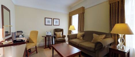 A hotel room with a wooden desk, two armchairs, a sofa, a small table, and a lamp. There are framed pictures on the wall and a window with curtains.