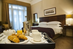 Una habitación de hotel con cama, mesita de noche con lámpara, ventana con cortinas y bandeja de desayuno con cruasanes, café y jugo.