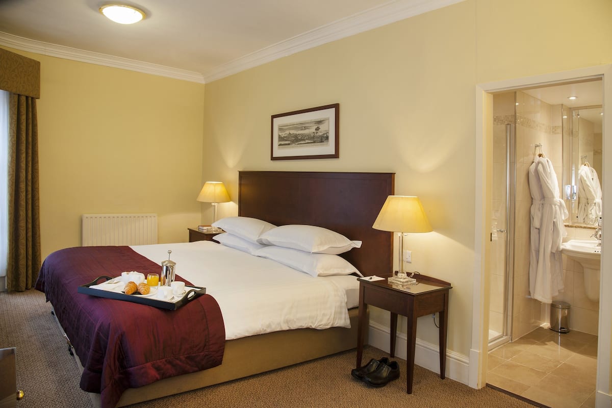 Une chambre d’hôtel avec un grand lit, deux lampes de chevet, un plateau avec des articles pour le petit-déjeuner, un tableau au mur et une salle de bain visible à travers une porte ouverte.