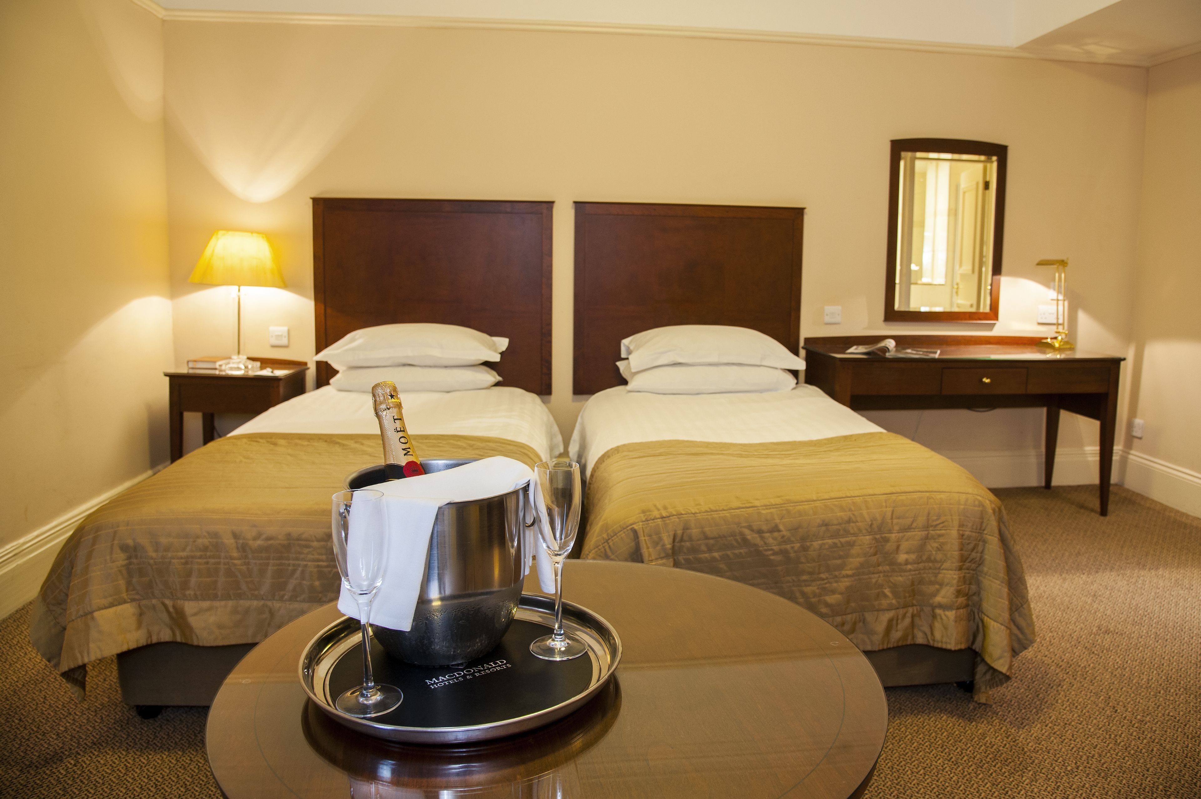 Een hotelkamer met twee bedden, een nachtkastje met lamp, een dienblad met champagnefles en glas, en een spiegel aan de muur.